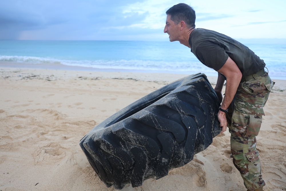 A man in military camouflage pants and a dark t-shirt is flipping a large heavy tire on a sandy beach. The activity appears to be a physical fitness or training exercise conducted in an outdoor beach environment. The ocean and cloudy sky are visible in the background.