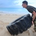 A man in military camouflage pants and a dark t-shirt is flipping a large heavy tire on a sandy beach. The activity appears to be a physical fitness or training exercise conducted in an outdoor beach environment. The ocean and cloudy sky are visible in the background.