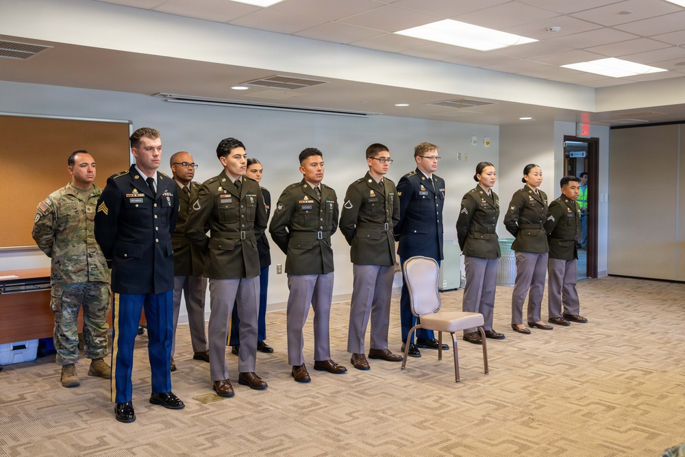 A group of approximately 11 military personnel are standing at attention in a line inside an indoor conference or meeting room. The individuals are wearing a mix of U.S. Army dress green uniforms, Army Service Uniforms (ASU), and one person in Army Combat Uniform (ACU/OCP). This appears to be a formal military ceremony or formation event.