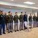 A group of approximately 11 military personnel are standing at attention in a line inside an indoor conference or meeting room. The individuals are wearing a mix of U.S. Army dress green uniforms, Army Service Uniforms (ASU), and one person in Army Combat Uniform (ACU/OCP). This appears to be a formal military ceremony or formation event.