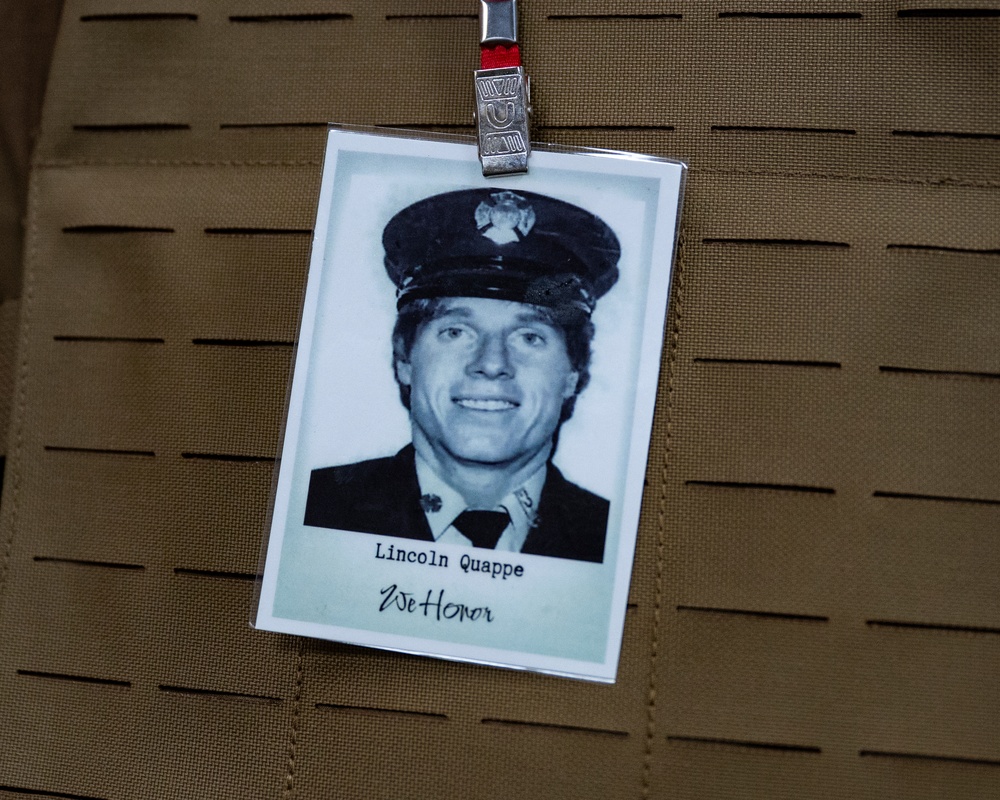 The image shows a memorial badge or tribute card clipped to what appears to be a tactical vest or MOLLE gear. The card features a black-and-white portrait photograph of a uniformed individual identified as 'Lincoln Quappe,' with the phrase 'We Honor' written below the name. This appears to be a commemorative tribute, likely honoring a fallen first responder.