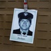 The image shows a memorial badge or tribute card clipped to what appears to be a tactical vest or MOLLE gear. The card features a black-and-white portrait photograph of a uniformed individual identified as 'Lincoln Quappe,' with the phrase 'We Honor' written below the name. This appears to be a commemorative tribute, likely honoring a fallen first responder.