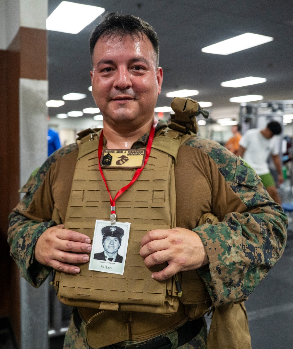 A U.S. Marine in woodland MARPAT camouflage uniform and tactical plate carrier vest is posing inside what appears to be a military fitness center or gym. He is displaying a badge/ID card on a red lanyard that contains a black-and-white photograph of another individual, likely a fallen service member being honored. The name patch on his plate carrier reads 'SANDOVAL, MIGUEL A' along with a USMC Eagle, Globe, and Anchor emblem.