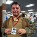 A U.S. Marine in woodland MARPAT camouflage uniform and tactical plate carrier vest is posing inside what appears to be a military fitness center or gym. He is displaying a badge/ID card on a red lanyard that contains a black-and-white photograph of another individual, likely a fallen service member being honored. The name patch on his plate carrier reads 'SANDOVAL, MIGUEL A' along with a USMC Eagle, Globe, and Anchor emblem.