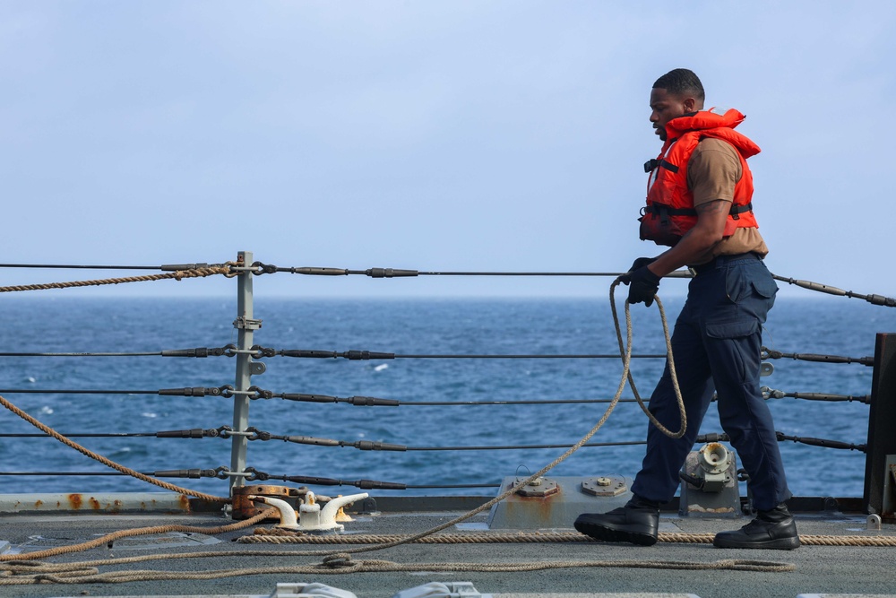 A sailor or naval personnel is working on the deck of a military or coast guard vessel at sea. The individual is handling a rope/line while wearing an orange life vest over a tan uniform with navy blue trousers. The open ocean is visible in the background beyond the ship's safety railing system.