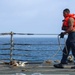 A sailor or naval personnel is working on the deck of a military or coast guard vessel at sea. The individual is handling a rope/line while wearing an orange life vest over a tan uniform with navy blue trousers. The open ocean is visible in the background beyond the ship's safety railing system.