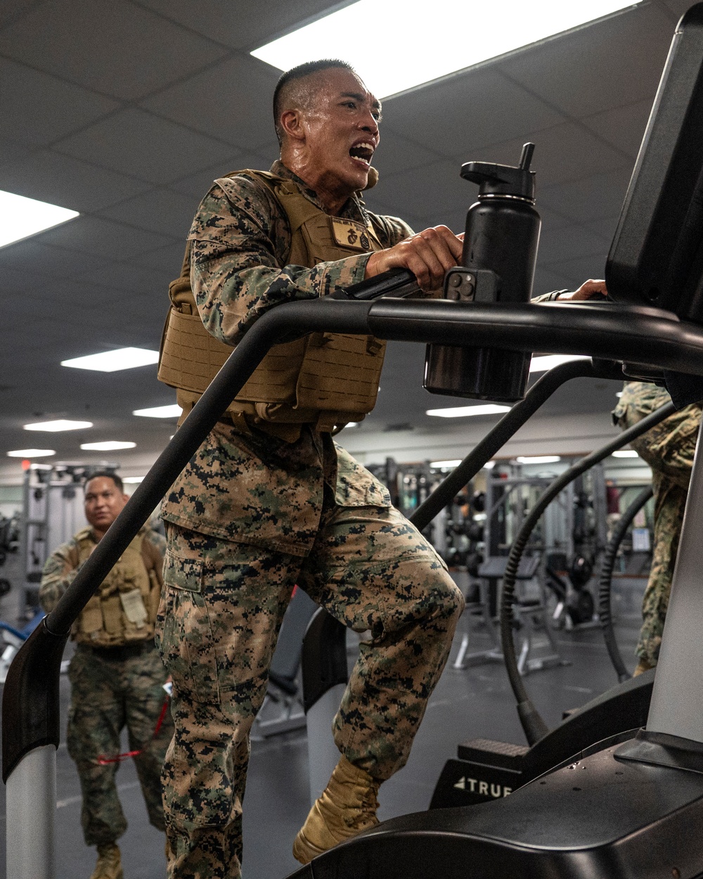 A U.S. Marine in full MARPAT camouflage uniform and tactical plate carrier vest is using a stair climber machine in a military fitness facility. The Marine appears to be exerting significant effort, with visible sweat and an open mouth expression indicating intense physical exertion. Additional Marines in similar uniforms are visible in the background using other gym equipment.