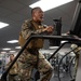 A U.S. Marine in full MARPAT camouflage uniform and tactical plate carrier vest is using a stair climber machine in a military fitness facility. The Marine appears to be exerting significant effort, with visible sweat and an open mouth expression indicating intense physical exertion. Additional Marines in similar uniforms are visible in the background using other gym equipment.
