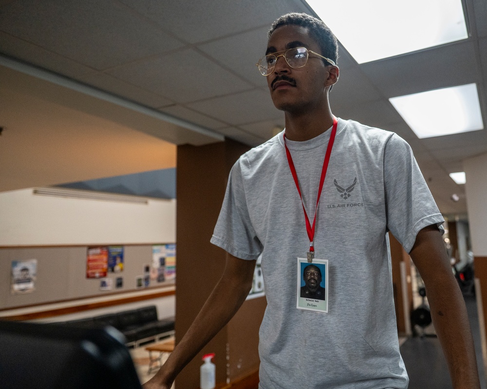 A young man wearing a U.S. Air Force t-shirt and gold-rimmed glasses stands in what appears to be an indoor facility, possibly a community center or government building. He is wearing a red lanyard with an ID badge around his neck. The background shows a common area with bulletin boards, seating, and fluorescent lighting.