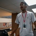 A young man wearing a U.S. Air Force t-shirt and gold-rimmed glasses stands in what appears to be an indoor facility, possibly a community center or government building. He is wearing a red lanyard with an ID badge around his neck. The background shows a common area with bulletin boards, seating, and fluorescent lighting.