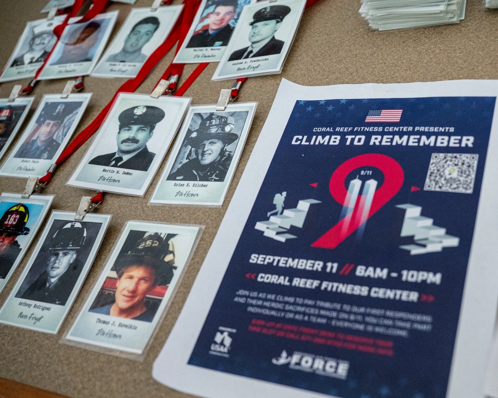 The image shows a memorial display for a '9/11 Climb to Remember' event hosted by Coral Reef Fitness Center. Multiple badge-style lanyards with black-and-white photographs of fallen first responders are laid out on a surface alongside an event flyer. The event is scheduled for September 11 from 8AM to 10PM at Coral Reef Fitness Center.