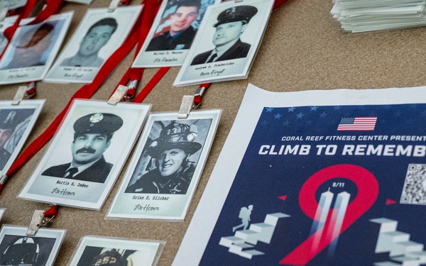 The image shows a memorial display for a '9/11 Climb to Remember' event hosted by Coral Reef Fitness Center. Multiple badge-style lanyards with black-and-white photographs of fallen first responders are laid out on a surface alongside an event flyer. The event is scheduled for September 11 from 8AM to 10PM at Coral Reef Fitness Center.
