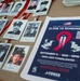 The image shows a memorial display for a '9/11 Climb to Remember' event hosted by Coral Reef Fitness Center. Multiple badge-style lanyards with black-and-white photographs of fallen first responders are laid out on a surface alongside an event flyer. The event is scheduled for September 11 from 8AM to 10PM at Coral Reef Fitness Center.