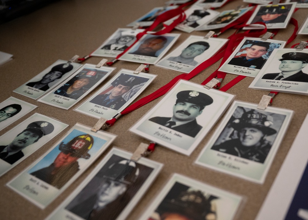 The image shows a memorial display featuring numerous ID badge-style photographs of firefighters and first responders laid out on a flat surface. Each photo is attached to a red lanyard and clipped with a small wooden clothespin, with handwritten names visible beneath each image. The display appears to be a tribute or remembrance collection honoring fallen or notable emergency service personnel.