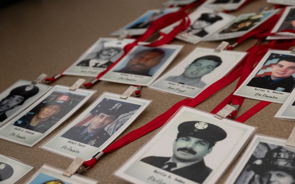 The image shows a memorial display featuring numerous ID badge-style photographs of firefighters and first responders laid out on a flat surface. Each photo is attached to a red lanyard and clipped with a small wooden clothespin, with handwritten names visible beneath each image. The display appears to be a tribute or remembrance collection honoring fallen or notable emergency service personnel.