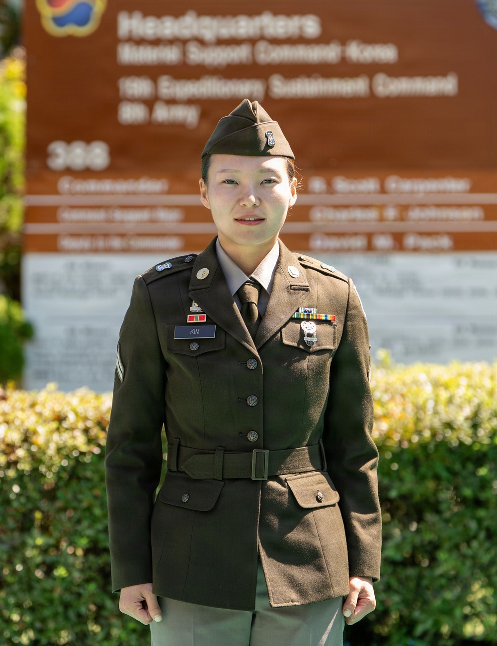 A U.S. Army soldier wearing the Army Service Uniform (ASU) in olive drab/green stands at attention in front of a military headquarters sign. The name tape reads 'KIM' and the soldier displays several ribbons, badges, and rank insignia. The background sign indicates 'Headquarters, Material Support Command Korea' and '19th Expeditionary Sustainment Command, 8th Army.'