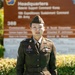 A U.S. Army soldier wearing the Army Service Uniform (ASU) in olive drab/green stands at attention in front of a military headquarters sign. The name tape reads 'KIM' and the soldier displays several ribbons, badges, and rank insignia. The background sign indicates 'Headquarters, Material Support Command Korea' and '19th Expeditionary Sustainment Command, 8th Army.'