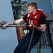 A young male sailor is photographed on the deck of a naval vessel, straining to pull a thick mooring or line-handling rope. He is wearing a red life vest over a black t-shirt, dark navy work trousers, and black boots. The image captures physical exertion during what appears to be a line-handling or mooring evolution.