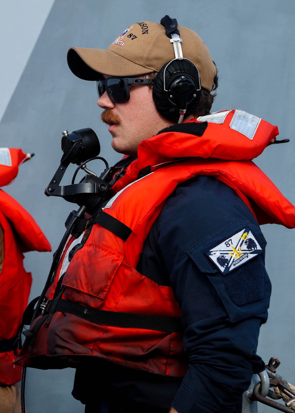 A naval or coast guard personnel member is shown in profile wearing an orange immersion/life vest over a dark blue uniform, with communication headset and a handheld microphone. A unit patch with the number '87' is visible on the left sleeve, and a tan cap with partial text reading '...SON 87' is worn on his head. The background suggests he is standing alongside a large gray vessel hull.