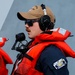 A naval or coast guard personnel member is shown in profile wearing an orange immersion/life vest over a dark blue uniform, with communication headset and a handheld microphone. A unit patch with the number '87' is visible on the left sleeve, and a tan cap with partial text reading '...SON 87' is worn on his head. The background suggests he is standing alongside a large gray vessel hull.