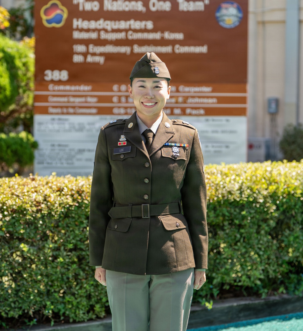 A U.S. Army soldier wearing the Army Service Uniform (ASU) in olive green stands smiling in front of a headquarters sign. The sign reads 'Two Nations, One Team' and identifies the location as Headquarters, Material Support Command-Korea, 19th Expeditionary Sustainment Command, 8th Army. The soldier displays a name tape reading 'LEE,' service ribbons, medals, and unit patches.