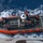 USCGC Bertholf conducts small boat operations in Timor Sea An aerial top-down view of a U.S. Coast Guard rigid-hull inflatable boat (RHIB) numbered 26576 on open ocean water. Four personnel in blue uniforms and orange life vests are visible aboard the vessel, which appears to be being recovered or deployed from a larger ship. The boat is orange with gray deck plating and is surrounded by white wake and deep blue ocean water.