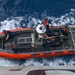 An aerial top-down view of a U.S. Coast Guard rigid-hull inflatable boat (RHIB) numbered 26576 on open ocean water. Four personnel in blue uniforms and orange life vests are visible aboard the vessel, which appears to be being recovered or deployed from a larger ship. The boat is orange with gray deck plating and is surrounded by white wake and deep blue ocean water.