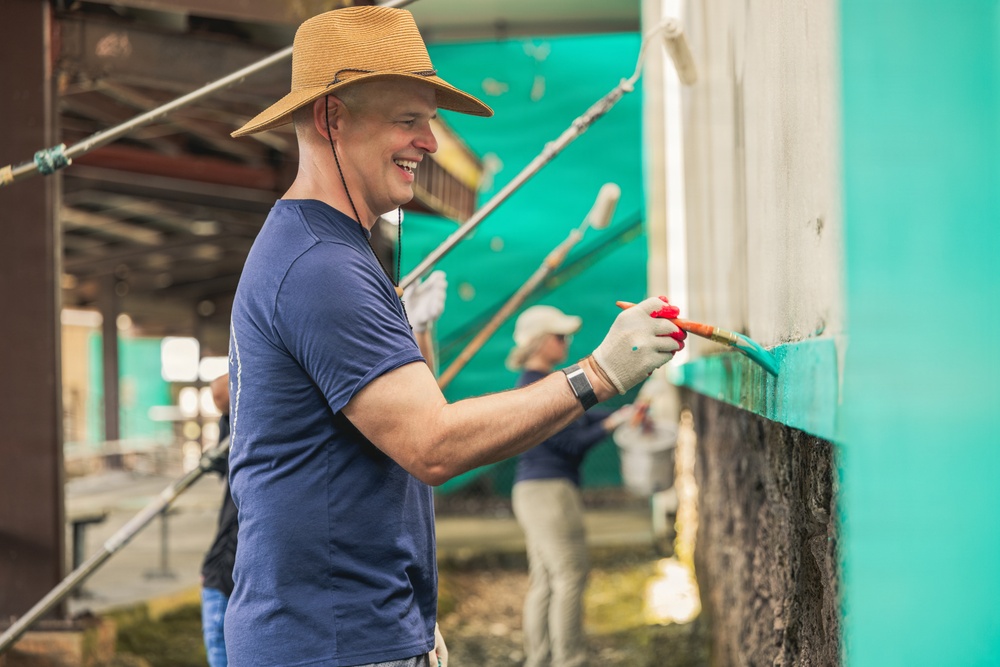 A smiling man wearing a straw hat and blue t-shirt is painting a wall with turquoise/teal paint using a brush. He is wearing work gloves and appears to be part of a community volunteer or renovation project. At least one other person is visible in the background performing similar painting work.