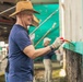 A smiling man wearing a straw hat and blue t-shirt is painting a wall with turquoise/teal paint using a brush. He is wearing work gloves and appears to be part of a community volunteer or renovation project. At least one other person is visible in the background performing similar painting work.