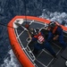 An aerial/overhead view of two U.S. Coast Guard personnel standing on the bow of an orange and gray rigid inflatable boat (RIB) numbered 26576. The boat is underway on open blue ocean water, with visible wake and chop surrounding the vessel. Both crew members are wearing blue uniforms, orange life vests, and black helmets.