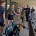 The image shows a ceremonial event at what appears to be a sports stadium or arena, featuring a large brass bell on a wooden stand as a central prop. A uniformed officer in a dark dress uniform with a campaign hat is rendering a salute, while civilians and military personnel in camouflage uniforms are present in the background. The setting suggests a military appreciation event or formal ceremony at a sporting venue.