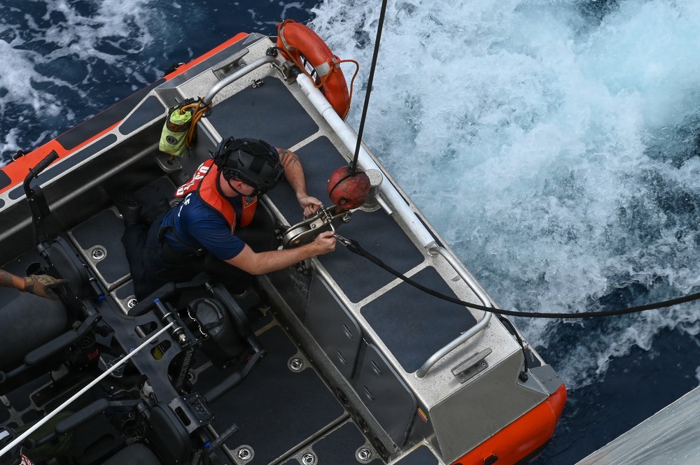 An aerial/overhead view of a Coast Guard rigid inflatable boat (RIB) on open ocean water. A crew member wearing an orange life vest and helmet is working on a rigging or hoisting mechanism at the stern of the vessel. The boat appears to be in motion, with white wake visible in the surrounding deep blue water.