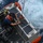 USCGC Bertholf conducts small boat operations in Timor Sea An aerial/overhead view of a Coast Guard rigid inflatable boat (RIB) on open ocean water. A crew member wearing an orange life vest and helmet is working on a rigging or hoisting mechanism at the stern of the vessel. The boat appears to be in motion, with white wake visible in the surrounding deep blue water.