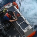 An aerial/overhead view of a Coast Guard rigid inflatable boat (RIB) on open ocean water. A crew member wearing an orange life vest and helmet is working on a rigging or hoisting mechanism at the stern of the vessel. The boat appears to be in motion, with white wake visible in the surrounding deep blue water.