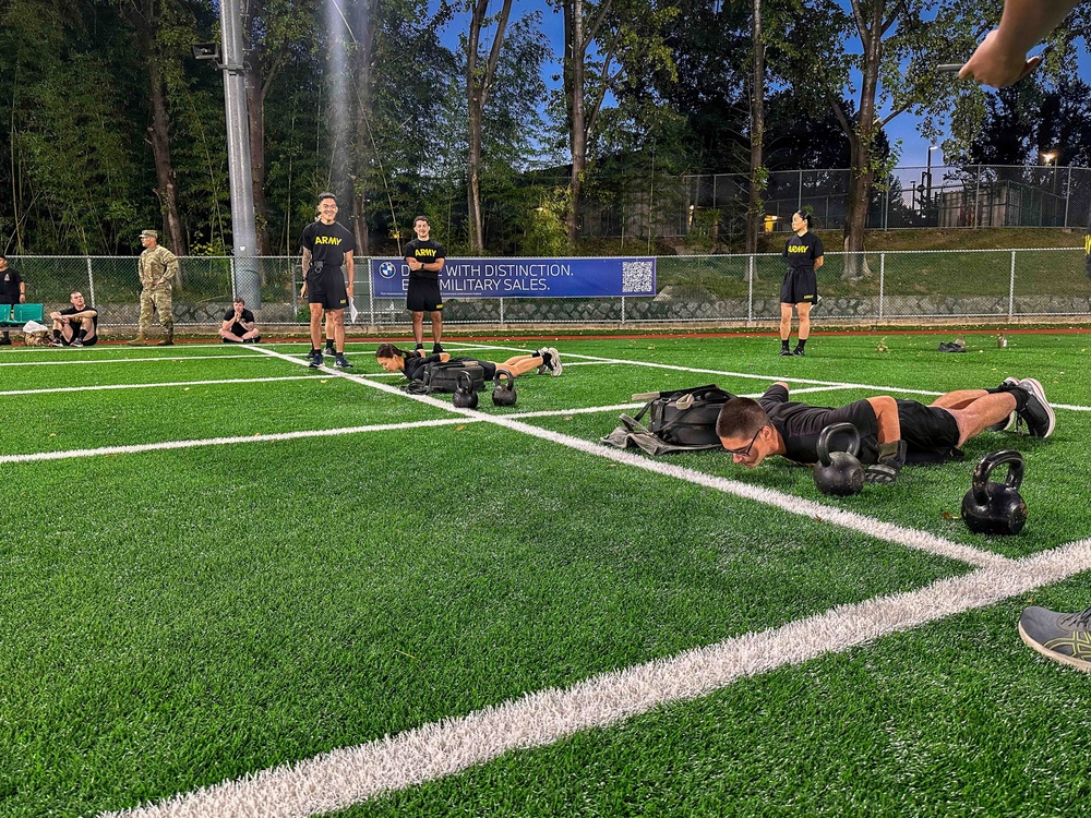A group of U.S. Army personnel are conducting a physical training session on an artificial turf athletic field. Several soldiers in black 'ARMY' branded PT uniforms are performing plank/push-up exercises with kettlebells and weighted sandbags, while others stand observing or supervising. A BMW military sales advertisement banner is visible along the perimeter fence.