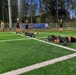 A group of U.S. Army personnel are conducting a physical training session on an artificial turf athletic field. Several soldiers in black 'ARMY' branded PT uniforms are performing plank/push-up exercises with kettlebells and weighted sandbags, while others stand observing or supervising. A BMW military sales advertisement banner is visible along the perimeter fence.