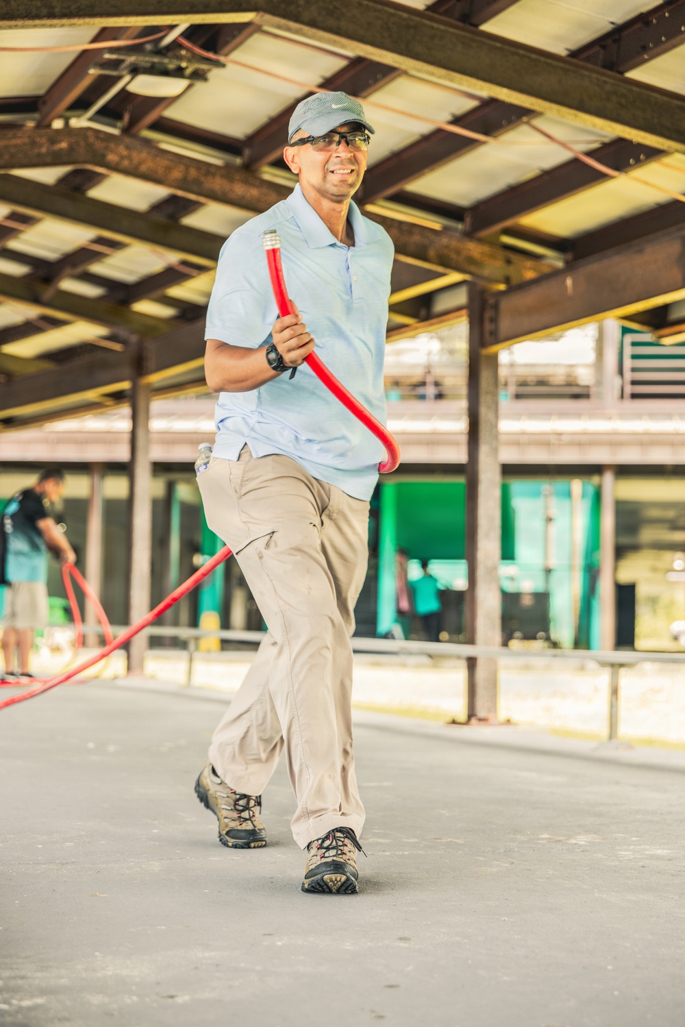 A man wearing a light blue polo shirt, khaki cargo pants, a gray cap, and safety glasses is walking while carrying a red hose or flexible pipe. He appears to be working in an industrial or construction setting with a steel-framed structure overhead. He is smiling and appears engaged in an active work task.