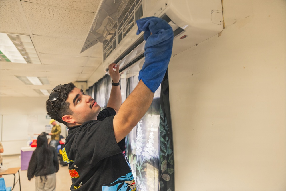 A young man is cleaning a wall-mounted mini-split air conditioning unit using a blue cloth or glove. He is wearing a black graphic hoodie with sunglasses resting on his head and a watch on his wrist. The setting appears to be an indoor community or classroom space with other people visible in the background.