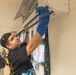 A young man is cleaning a wall-mounted mini-split air conditioning unit using a blue cloth or glove. He is wearing a black graphic hoodie with sunglasses resting on his head and a watch on his wrist. The setting appears to be an indoor community or classroom space with other people visible in the background.