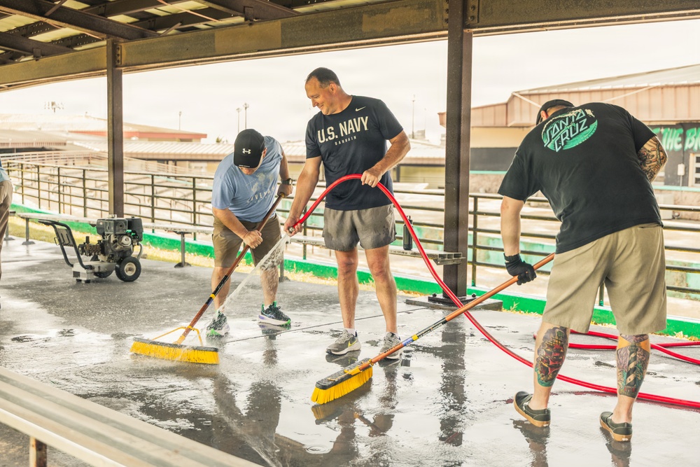 Three men are cleaning a wet concrete floor under a covered outdoor structure, using push brooms and a water hose connected to a pressure washer. One man is wearing a 'U.S. Navy Bahrain' shirt, another wears an Under Armour cap and blue shirt, and the third wears a Santa Cruz branded black shirt with visible tattoos on his arms. The setting appears to be a fairground or similar outdoor facility.