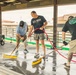 Three men are cleaning a wet concrete floor under a covered outdoor structure, using push brooms and a water hose connected to a pressure washer. One man is wearing a 'U.S. Navy Bahrain' shirt, another wears an Under Armour cap and blue shirt, and the third wears a Santa Cruz branded black shirt with visible tattoos on his arms. The setting appears to be a fairground or similar outdoor facility.