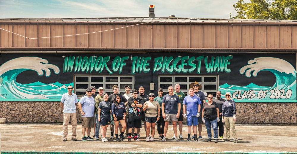 A group of approximately 18-20 people pose in front of a building featuring a large mural with ocean waves and the text 'IN HONOR OF THE BIGGEST WAVE - CLASS OF 2020.' Several individuals appear to be wearing U.S. Navy-affiliated clothing, suggesting a military community service or volunteer event. The mural appears freshly painted, indicating the group may have participated in its creation.