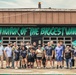 A group of approximately 18-20 people pose in front of a building featuring a large mural with ocean waves and the text 'IN HONOR OF THE BIGGEST WAVE - CLASS OF 2020.' Several individuals appear to be wearing U.S. Navy-affiliated clothing, suggesting a military community service or volunteer event. The mural appears freshly painted, indicating the group may have participated in its creation.