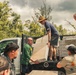 A group of people are unloading building materials from the flatbed of a white pickup truck in an outdoor setting with lush green vegetation in the background. The individuals appear to be engaged in a community service or disaster relief effort, handling large flat panels and stacked boxes labeled 'SHENGLUDA CEILING'. At least one person visible on the right is wearing what appears to be a military or uniformed shirt with insignia patches.