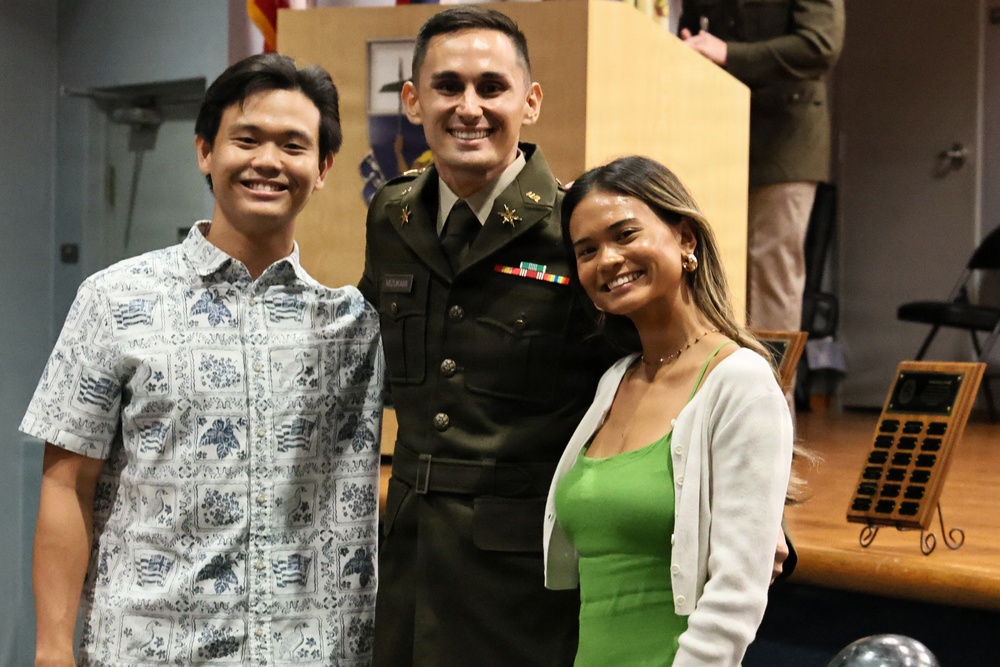 Three smiling individuals pose together at what appears to be a military ceremony or formal event. The person in the center is wearing a U.S. Army dress green uniform with rank insignia, ribbons, and a name tag. They are flanked by two civilians — a young man in a Hawaiian-style shirt on the left and a young woman in a green top with a white cardigan on the right.