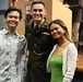 Three smiling individuals pose together at what appears to be a military ceremony or formal event. The person in the center is wearing a U.S. Army dress green uniform with rank insignia, ribbons, and a name tag. They are flanked by two civilians — a young man in a Hawaiian-style shirt on the left and a young woman in a green top with a white cardigan on the right.