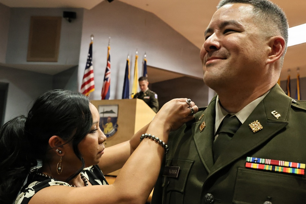 A woman is pinning an insignia or device onto the collar/chest area of a U.S. Army officer in dress green uniform during what appears to be a promotion or award ceremony. The officer is smiling and wearing multiple service ribbons and branch insignia. The ceremony is taking place in a formal indoor venue with multiple flags displayed in the background.