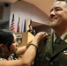 A woman is pinning an insignia or device onto the collar/chest area of a U.S. Army officer in dress green uniform during what appears to be a promotion or award ceremony. The officer is smiling and wearing multiple service ribbons and branch insignia. The ceremony is taking place in a formal indoor venue with multiple flags displayed in the background.