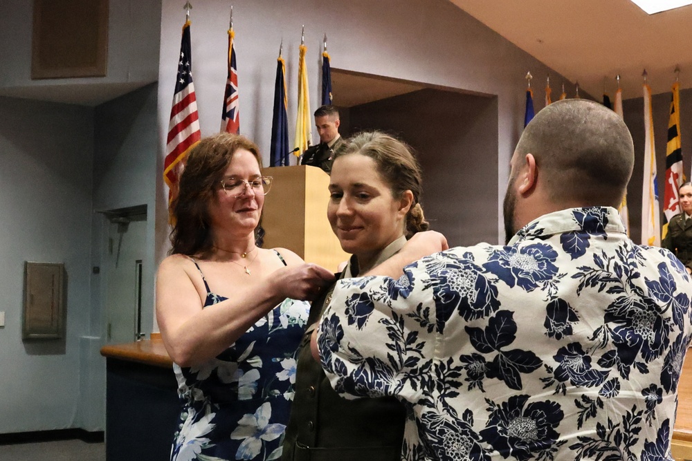 A military promotion or commissioning ceremony is taking place indoors, where a woman in military uniform is having insignia or a pin affixed to her uniform by two civilians — an older woman on the left and a man on the right. Multiple national and organizational flags are displayed in the background, and a uniformed officer stands at a podium. The setting appears to be an official military ceremony hall.