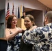 A military promotion or commissioning ceremony is taking place indoors, where a woman in military uniform is having insignia or a pin affixed to her uniform by two civilians — an older woman on the left and a man on the right. Multiple national and organizational flags are displayed in the background, and a uniformed officer stands at a podium. The setting appears to be an official military ceremony hall.